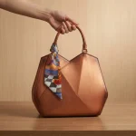 Hand holding a bronze geometric leather handbag with an artistic wearable art silk scarf knotted around the handle base.
