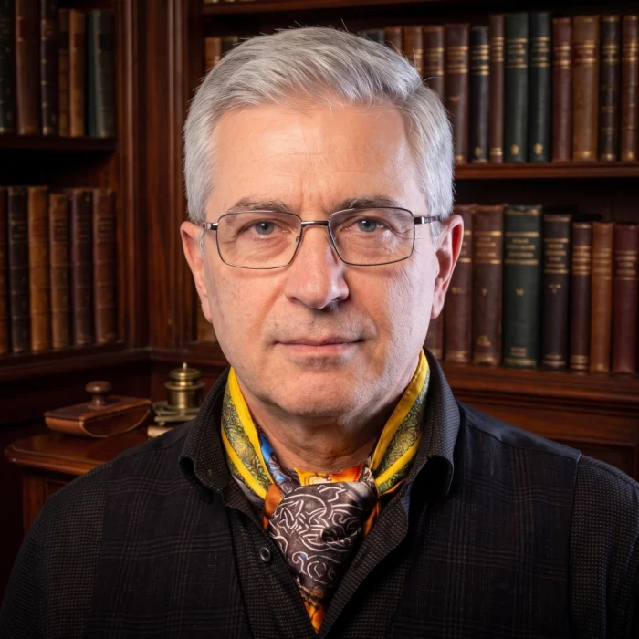 Mature man with glasses wearing a patterned wearable art silk scarf tied as an ascot with a dark collared shirt in front of a wooden bookshelf.