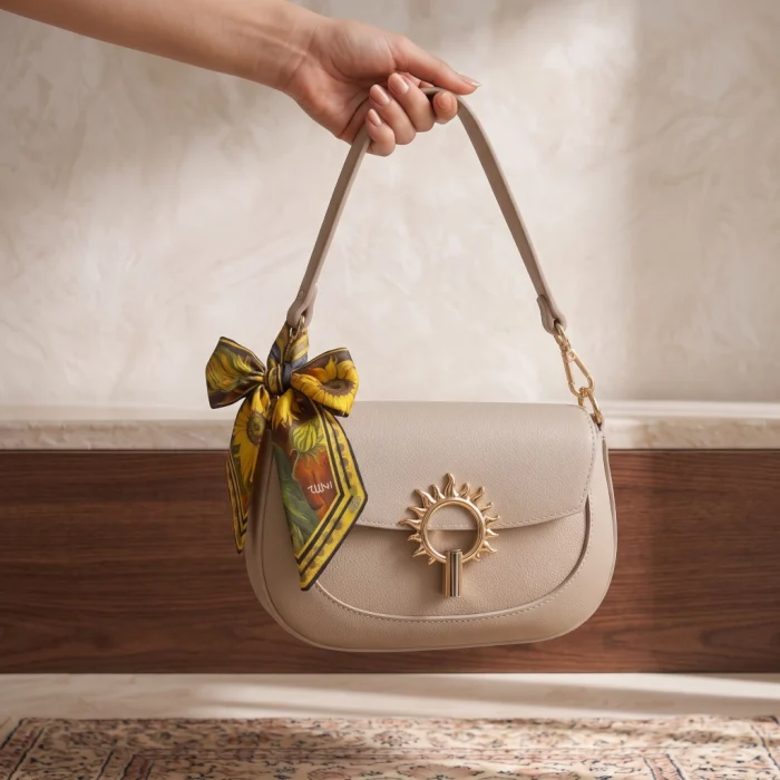Hand holding a beige leather handbag with a gold sun clasp and a sunflower-print wearable art silk scarf tied in a bow to the strap.